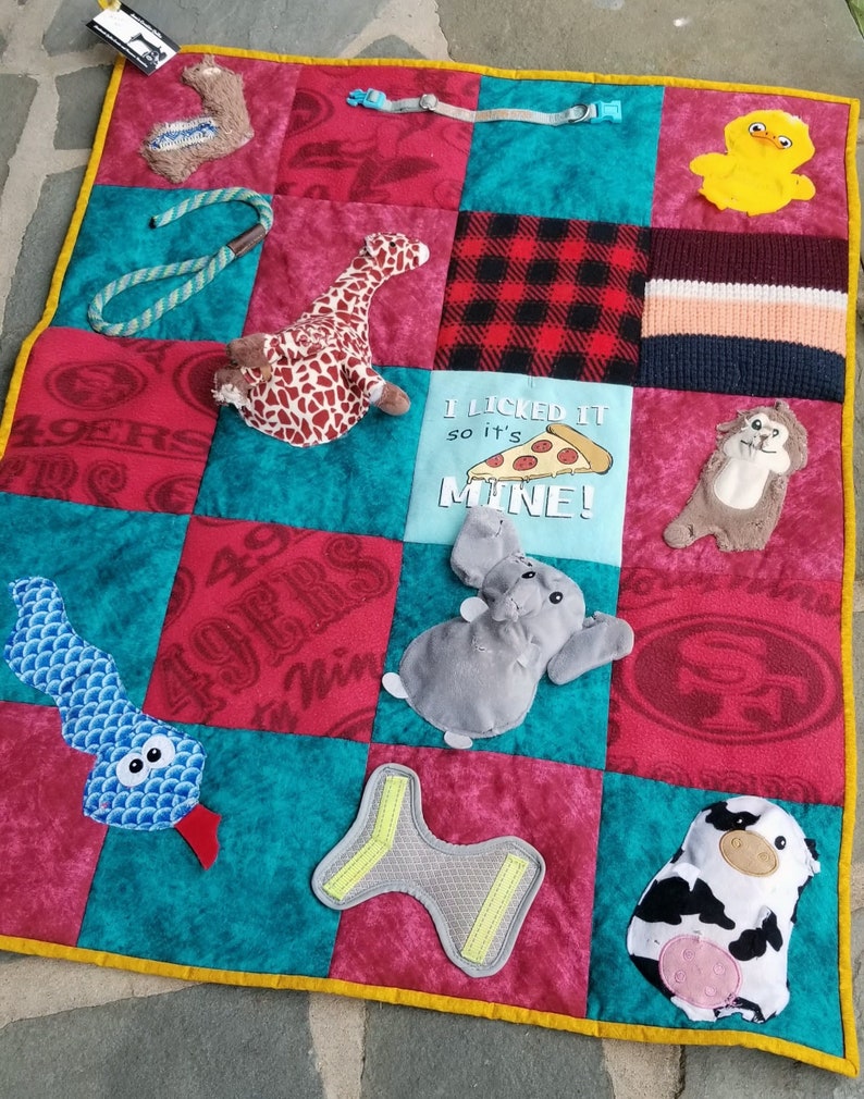 Pu&ograve; includere: Un tappetino da gioco trapuntato multicolore per cani con diverse trame e forme. Il tappetino presenta un tessuto rosso, bianco e blu con il testo "I licked it so it's mine!" e una grafica a fetta di pizza. Altre caratteristiche includono un serpente, una giraffa, un elefante, una mucca, un'anatra e un giocattolo a forma di osso.
