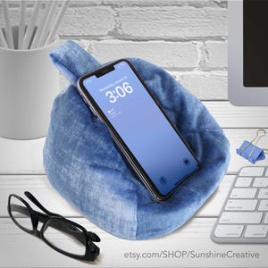 May include: A blue fabric phone holder with a handle, holding a smartphone displaying the time 3:06. The holder is on a white surface with a keyboard, glasses, and a pen holder in the background. The Etsy shop name is visible.