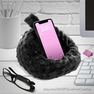May include: A black, plush phone holder with a textured surface cradles a smartphone with a pink screen displaying the time 3:06. The holder sits on a white surface, with a pair of black glasses and a keyboard nearby.