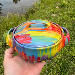 May include: A round ceramic casserole dish with a lid, featuring a vibrant, multi-colored marbled glaze in shades of red, yellow, blue, and green. The dish has two side handles and three small steam vent holes on the top of the lid.