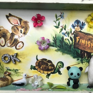 Puede incluir: Un diorama en miniatura con un tema de bosque caprichoso. Presenta un conejito marrón, un pájaro azul, una tortuga, un oso panda, un pingüino y un pato. También hay flores y un cartel que dice "Finish".