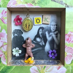 May include: A framed mixed media artwork with a religious pendant, a baby doll, and a glitter background. The artwork also includes a black and white photo of two women, a pink flower, and a purple flower. The letters "OK" are spelled out with wooden blocks.