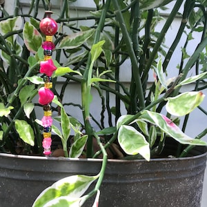 May include: A pink and gold beaded garland with a red glass bead at the top. The garland is hanging from a plant in a black metal pot.