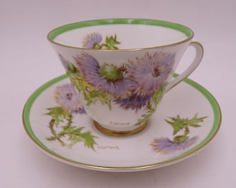 May include: A white teacup and saucer with a green rim and a floral design of purple thistles. The saucer has the word "Scotland" written on it.