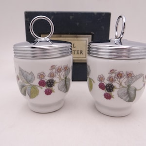 May include: Two white ceramic egg coddlers with silver lids and a blackberry floral design. The coddlers are in a blue box with the word "Coddler" printed on it.