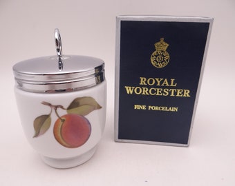 Könnte beinhalten: Eine weiße Porzellan-Eierbecher mit einem silbernen Deckel und einem Pfirsich-Design. Der Eierbecher befindet sich in seiner Originalverpackung, die blau ist mit goldenen Schriftzug "Royal Worcester Fine Porcelain".