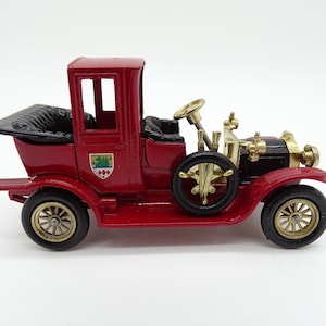 Op de afbeelding: Een rode speelgoedauto met zwarte stoelen en gouden accenten. De auto heeft een zwart dak en een gouden stuurwiel. De auto is een vintage model en heeft een wapenschild aan de zijkant.
