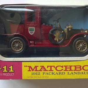 May include: A red 1912 Packard Landaulet die-cast model car by Matchbox, Lesney product Y-11, displayed in its original yellow and pink cardboard box with a clear plastic window. The vintage-style vehicle features gold-colored wheels and trim.