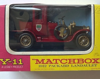 Puede incluir: Un coche de juguete modelo 1912 Packard Landaulet de color rojo de Matchbox, producto Lesney Y-11, presentado en su caja de cartón original amarilla y rosa con ventana de plástico. El vehículo cuenta con ruedas y detalles en color dorado.