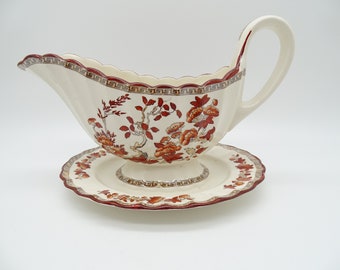 May include: A white gravy boat with a red and gold floral pattern and a matching saucer. The gravy boat has a scalloped edge and a handle.