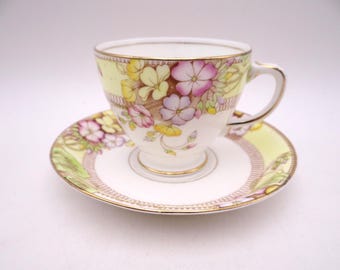 May include: A white porcelain teacup and saucer set featuring a delicate floral pattern with pink, yellow, and purple flowers. The design includes a pale yellow background, intricate brown lattice detailing, and gold trim on the edges.