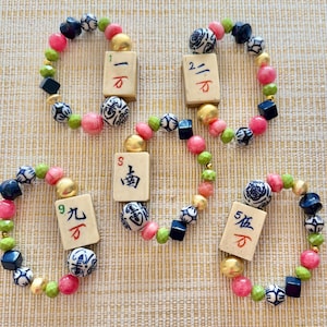 Puede incluir: Cuatro pulseras de cuentas con cuentas rosas, verdes, doradas, azul marino y blancas. Cada pulsera presenta una baldosa rectangular con caracteres chinos. Las pulseras est&aacute;n dispuestas sobre una superficie clara y texturizada.