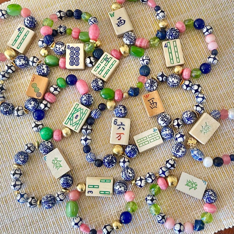 Puede incluir: Una colecci&oacute;n de pulseras de cuentas con una variedad de cuentas coloridas en rosa, verde, azul y dorado. Cada pulsera presenta peque&ntilde;as fichas de Mahjong rectangulares con caracteres y s&iacute;mbolos chinos. Las pulseras est&aacute;n dispuestas sobre una superficie de color beige claro.