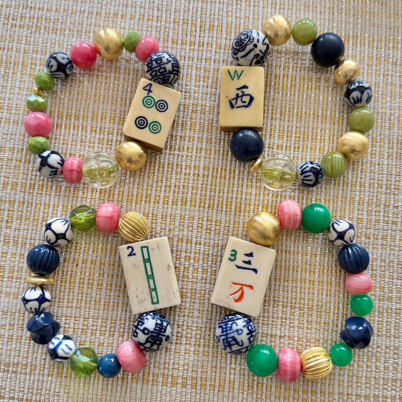 Puede incluir: Cuatro pulseras de cuentas con una variedad de cuentas coloridas, que incluyen esferas rosas, verdes, doradas y estampadas. Cada pulsera presenta una ficha de Mahjong como pieza central. Las fichas muestran n&uacute;meros y caracteres chinos.