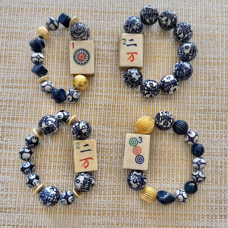 Puede incluir: Cuatro pulseras hechas a mano con cuentas estampadas en azul y blanco, cuentas azul oscuro y detalles dorados. Cada pulsera incorpora una ficha de Mahjong, a&ntilde;adiendo un toque &uacute;nico al dise&ntilde;o.