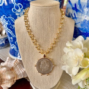 Può includere: Una collana a catena color oro con un ciondolo circolare che presenta una moneta color argento al centro. La moneta mostra un ritratto di profilo, il testo E PLURIBUS UNUM e l'anno 1886. Il ciondolo è fissato a una spessa catena a maglie dorate.
