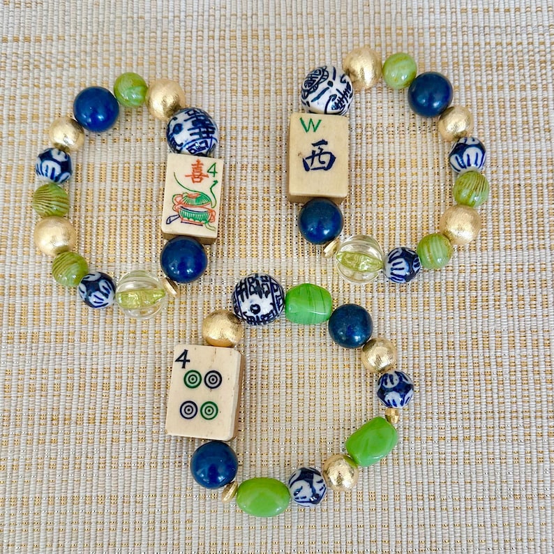 Puede incluir: Tres pulseras de cuentas con cuentas doradas, azules, verdes y blancas. Cada pulsera presenta una ficha de Mahjong. Las fichas muestran caracteres y n&uacute;meros chinos.