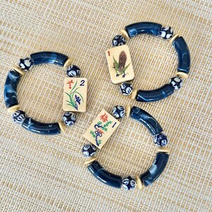 Puede incluir: Tres pulseras con cuentas azules y blancas y fichas de Mahjong. Las pulseras presentan cuentas curvas de color azul oscuro, cuentas estampadas y espaciadores color crema. Cada pulsera tiene una ficha diferente con ilustraciones coloridas.
