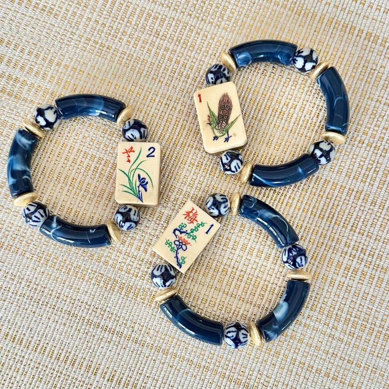 Puede incluir: Tres pulseras con cuentas azules y blancas y fichas de Mahjong. Las pulseras presentan cuentas curvas de color azul oscuro, cuentas estampadas y espaciadores color crema. Cada pulsera tiene una ficha diferente con ilustraciones coloridas.