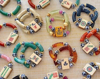 Puede incluir: Una colección de pulseras circulares que presentan fichas de mahjong individuales como piezas centrales. Cada pulsera está compuesta por cuentas de acrílico o resina de colores, pequeños espaciadores de cerámica azul y blanca.