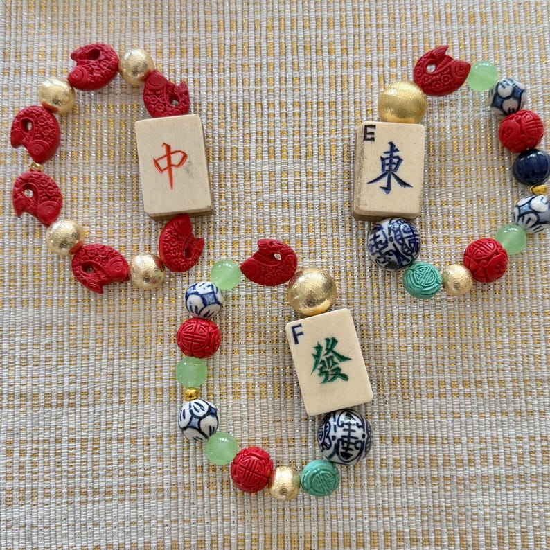 Puede incluir: Tres pulseras de cuentas con tem&aacute;tica Mahjong. Cada pulsera presenta cuentas rojas, doradas y verdes, junto con fichas de Mahjong de color marfil con caracteres chinos. Las pulseras est&aacute;n dispuestas sobre una superficie beige texturizada.