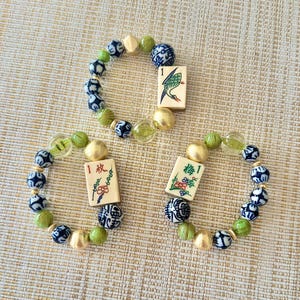 Puede incluir: Tres pulseras de cuentas con una mezcla de cuentas verdes, doradas y azules. Cada pulsera presenta una peque&ntilde;a baldosa rectangular con un dise&ntilde;o decorativo. Las pulseras est&aacute;n dispuestas sobre una superficie beige texturizada.