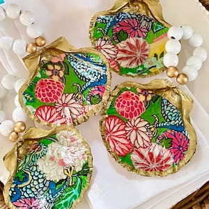 Pode incluir: Quatro conchas douradas com fundo verde e padrões florais e de pássaros coloridos. As conchas estão dispostas em círculo sobre uma superfície branca.