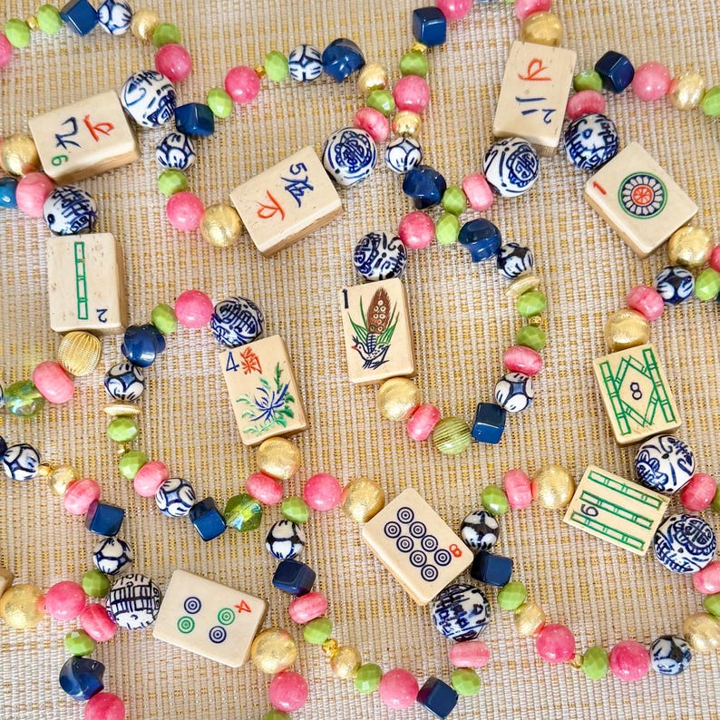 Puede incluir: Collares de cuentas con fichas de Mahjong de color marfil, cuentas rosas, verdes, doradas y azules. Las fichas muestran s&iacute;mbolos y caracteres chinos, creando un accesorio &uacute;nico y l&uacute;dico.