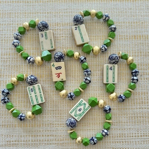 Puede incluir: Cinco pulseras de cuentas con una ficha de Mahjong en el centro. Las pulseras presentan cuentas verdes, doradas y azules. Las fichas de Mahjong tienen n&uacute;meros y s&iacute;mbolos. Las pulseras est&aacute;n sobre una superficie beige.