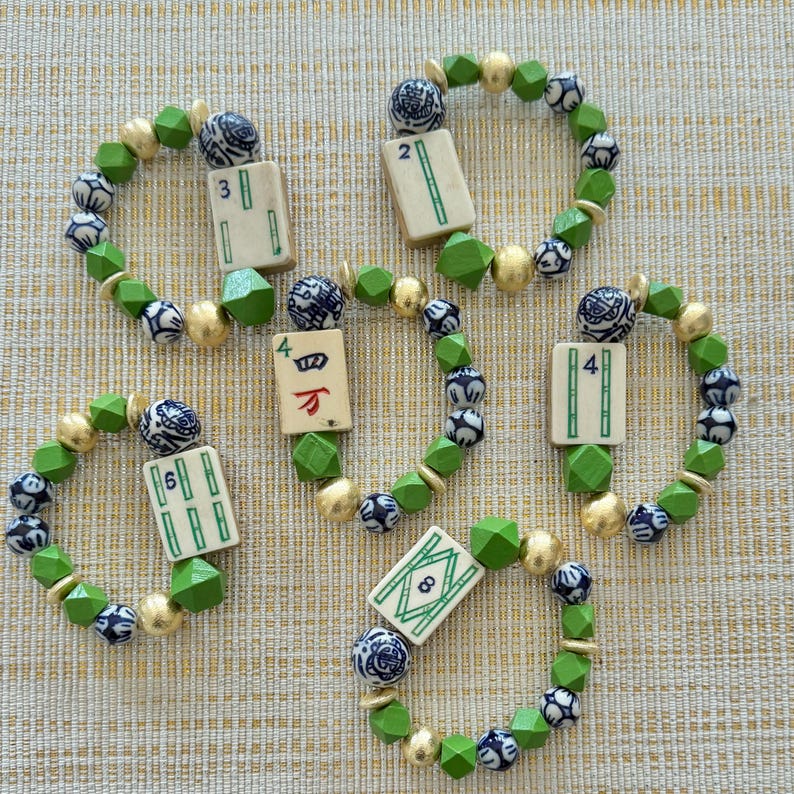 Puede incluir: Cinco pulseras de cuentas con una ficha de Mahjong en el centro. Las pulseras presentan cuentas verdes, doradas y azules. Las fichas de Mahjong tienen n&uacute;meros y s&iacute;mbolos. Las pulseras est&aacute;n sobre una superficie beige.