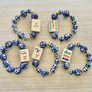 Puede incluir: Cuatro pulseras con cuentas estampadas en azul y blanco y espaciadores dorados. Cada pulsera tiene una ficha de Mahjong. Las fichas tienen varios s&iacute;mbolos y n&uacute;meros. Las pulseras est&aacute;n sobre una superficie beige.