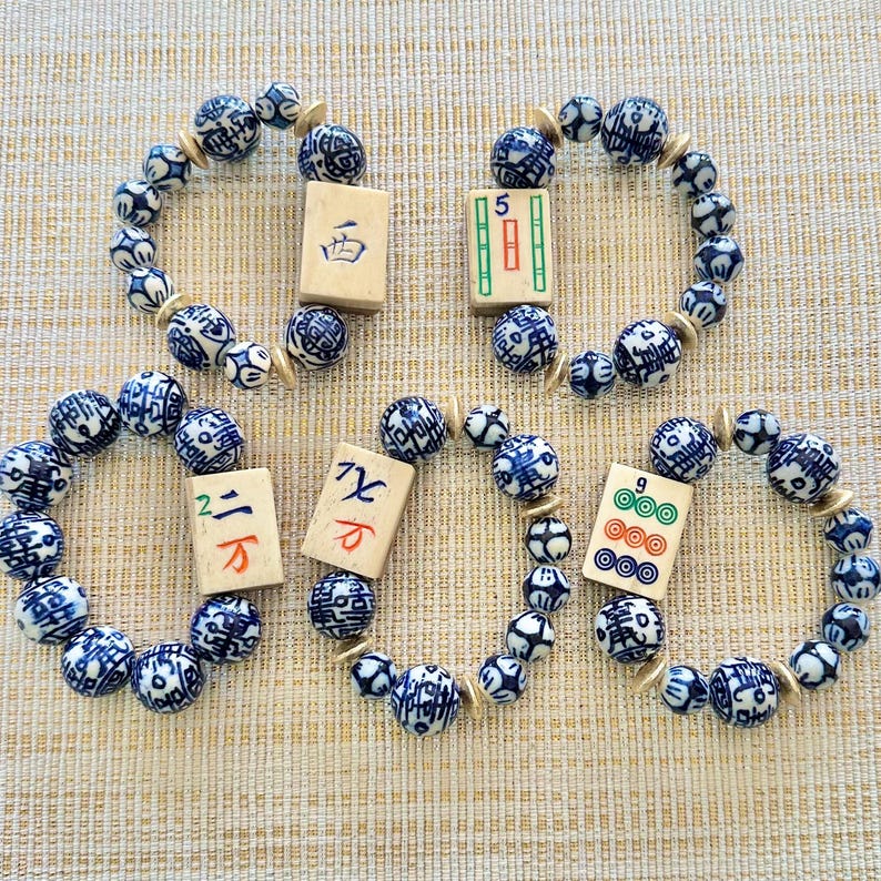 Puede incluir: Cuatro pulseras con cuentas estampadas en azul y blanco y espaciadores dorados. Cada pulsera tiene una ficha de Mahjong. Las fichas tienen varios s&iacute;mbolos y n&uacute;meros. Las pulseras est&aacute;n sobre una superficie beige.