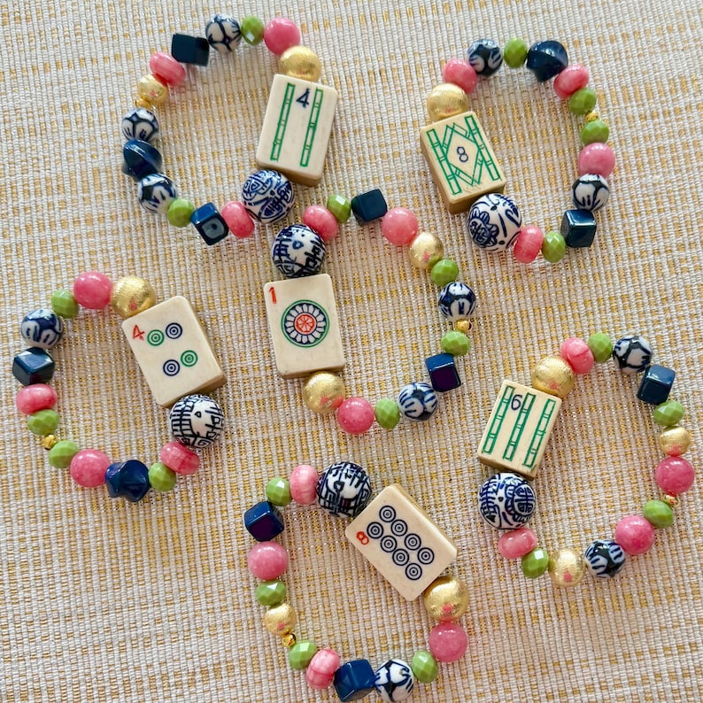 Puede incluir: Cinco pulseras de cuentas con una pieza central de azulejo de Mahjong. Las pulseras presentan cuentas rosas, verdes, doradas, azules y blancas. Las fichas de Mahjong tienen marcas verdes y negras. Las pulseras est&aacute;n sobre una superficie clara y texturizada.