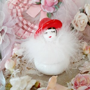 May include: A white ceramic figurine of a woman wearing a red hat and a fluffy white feather boa. The figurine has painted facial features and is surrounded by pink roses and lace.