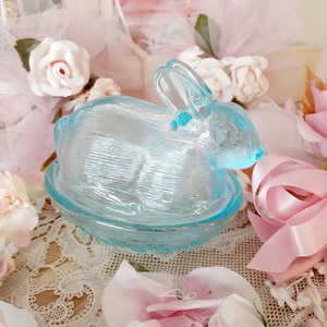 May include: A light blue glass bunny figurine sitting on a matching dish. The bunny is surrounded by pink and white flowers and lace. The figurine is a decorative item.