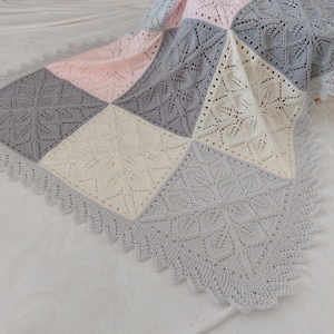 May include: A crocheted baby blanket with a square pattern in shades of pink, cream, and gray. The blanket has a delicate lace border.