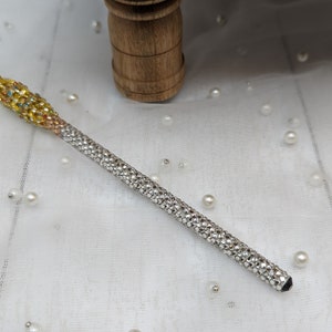 May include: A decorative wand with a gradient design. The top section is gold and adorned with colorful rhinestones, transitioning to a silver, crystal-covered shaft. The wand is set against a white, pearl-studded background.