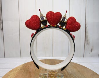 May include: A black headband with a white interior, adorned with three large, glittery red heart shapes. The headband also features smaller black heart accents and decorative elements, creating a festive and eye-catching accessory.