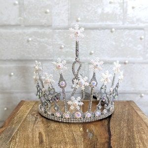 May include: A silver crown with white snowflake accents and iridescent rhinestones. The crown is on a wooden surface.