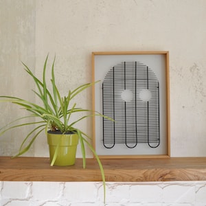 Puede incluir: Una escultura de alambre negro enmarcada de un fantasma con dos ojos redondos, colgada en una pared sobre un estante de madera con una planta verde en maceta. El estante está sobre una pared de azulejos blancos con un grifo plateado.