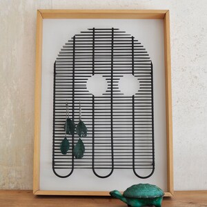 Op de afbeelding: Een zwart metalen draadframe in de vorm van een spook met twee ooggaten, hangend aan een muur in een houten frame. Het spook is versierd met groene, blaadjesvormige oorbellen. Een klein groen schildpadvormig sieradenkistje staat op een houten plank onder het frame.