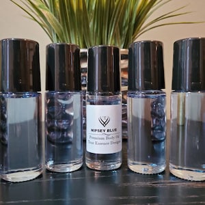 Può includere: Cinque flaconi neri a rullo contenenti un liquido trasparente. Il flacone al centro ha un'etichetta con la scritta "Nipsey Blue Premium Body Oil True Essence Design".