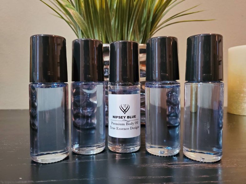 May include: Five black roll-on bottles containing clear liquid. The central bottle has a label reading "Nipsey Blue Premium Body Oil True Essence Design".