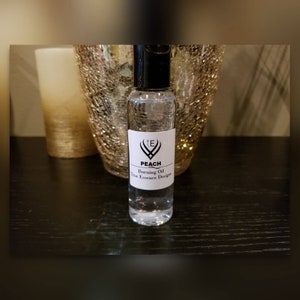 May include: A clear glass bottle with a black cap and a white label. The label says "TE Peach Burning Oil True Essence Designs".