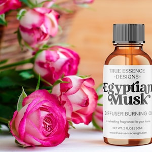 Może przedstawiać: Brązowa szklana butelka z czarną zakrętką i białą etykietą z napisem "TRUE ESSENCE -DESIGNS- Egyptian Musk DIFFUSER|BURNING OIL a refreshing fragrance for your home NET WT. 2 FLOZ | 60ML www.trueessencedesigns.com". Butelka stoi na białym tle z różowymi różami w tle.