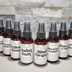 May include: A collection of eight brown glass spray bottles with black spray nozzles and clear caps. Each bottle has a white label with a different scent name, such as "Passion Kisses" and "Pink Peony". The bottles are labeled "Room Car Spray".
