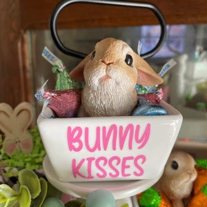May include: A white ceramic dish with the words "Bunny Kisses" in pink lettering. A small brown bunny figurine is sitting inside the dish, surrounded by colorful foil-wrapped chocolate candies.