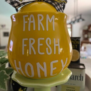 May include: A yellow ceramic honey pot with the words "FARM FRESH HONEY" in white. The pot has a lid and is decorated with a bee-themed ribbon. It sits on a light green pedestal.