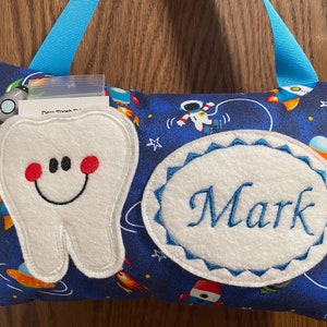May include: A blue tooth pillow with a white tooth and a white oval with the name "Mark" embroidered in blue. The pillow has a space theme with rockets, stars, and an astronaut.