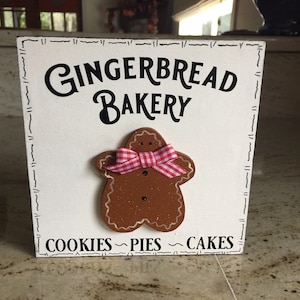 May include: White wooden sign with black text that reads "Gingerbread Bakery" and a gingerbread man illustration with a red and white bow. The sign also says "Cookies ~ Pies ~ Cakes".